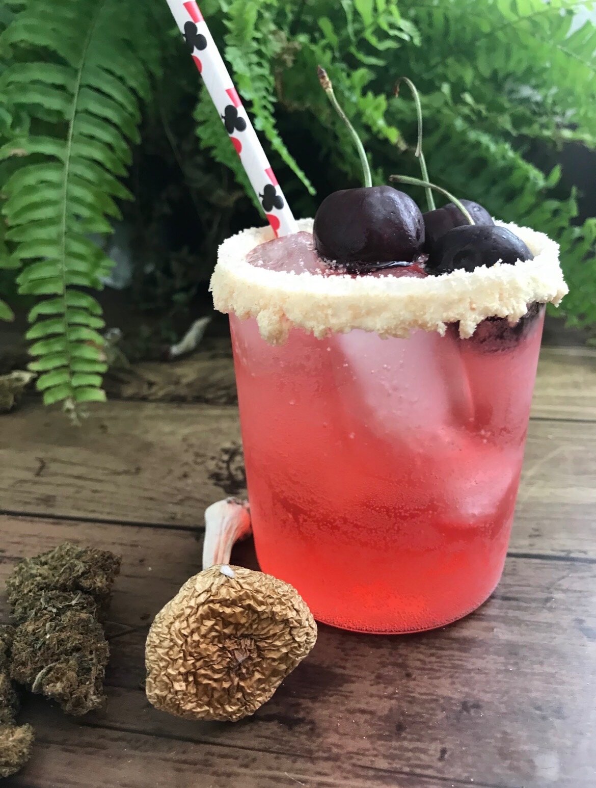 Pink cherry cocktail with sugared rim, ice, and dark cherries beside dried mushrooms on wooden table.