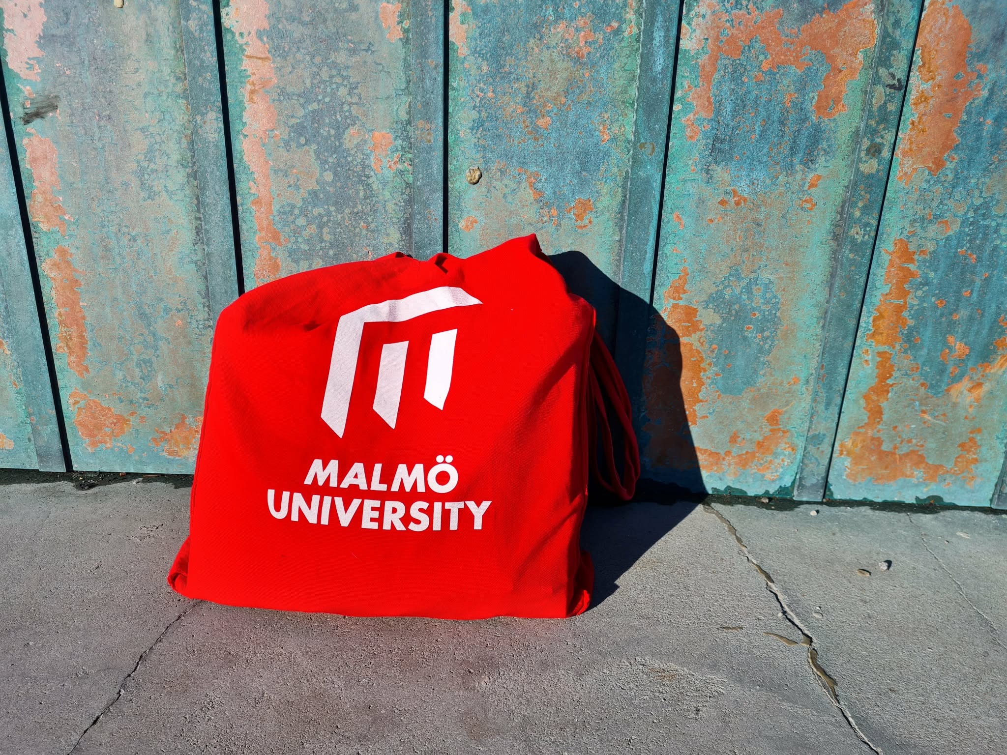 Red Malmö University tote bag against a weathered turquoise metal wall.