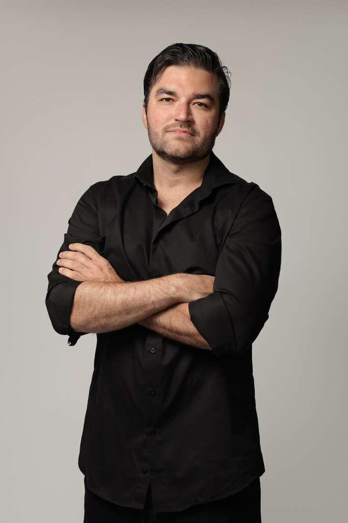 Man in black button-down shirt posing with arms crossed against a neutral studio background.