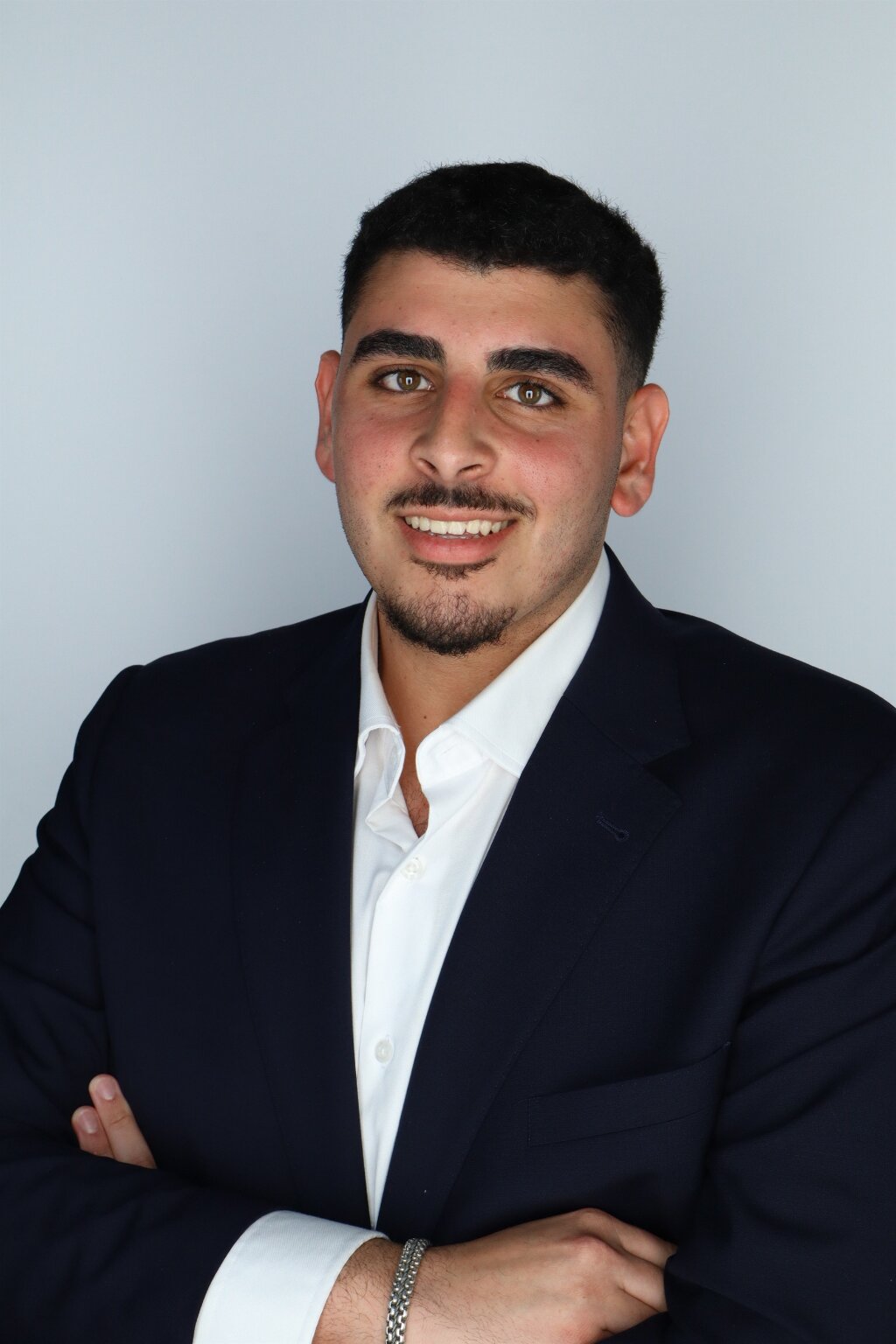 Smiling Eli Dabach in navy suit and white shirt with arms crossed against gray background.