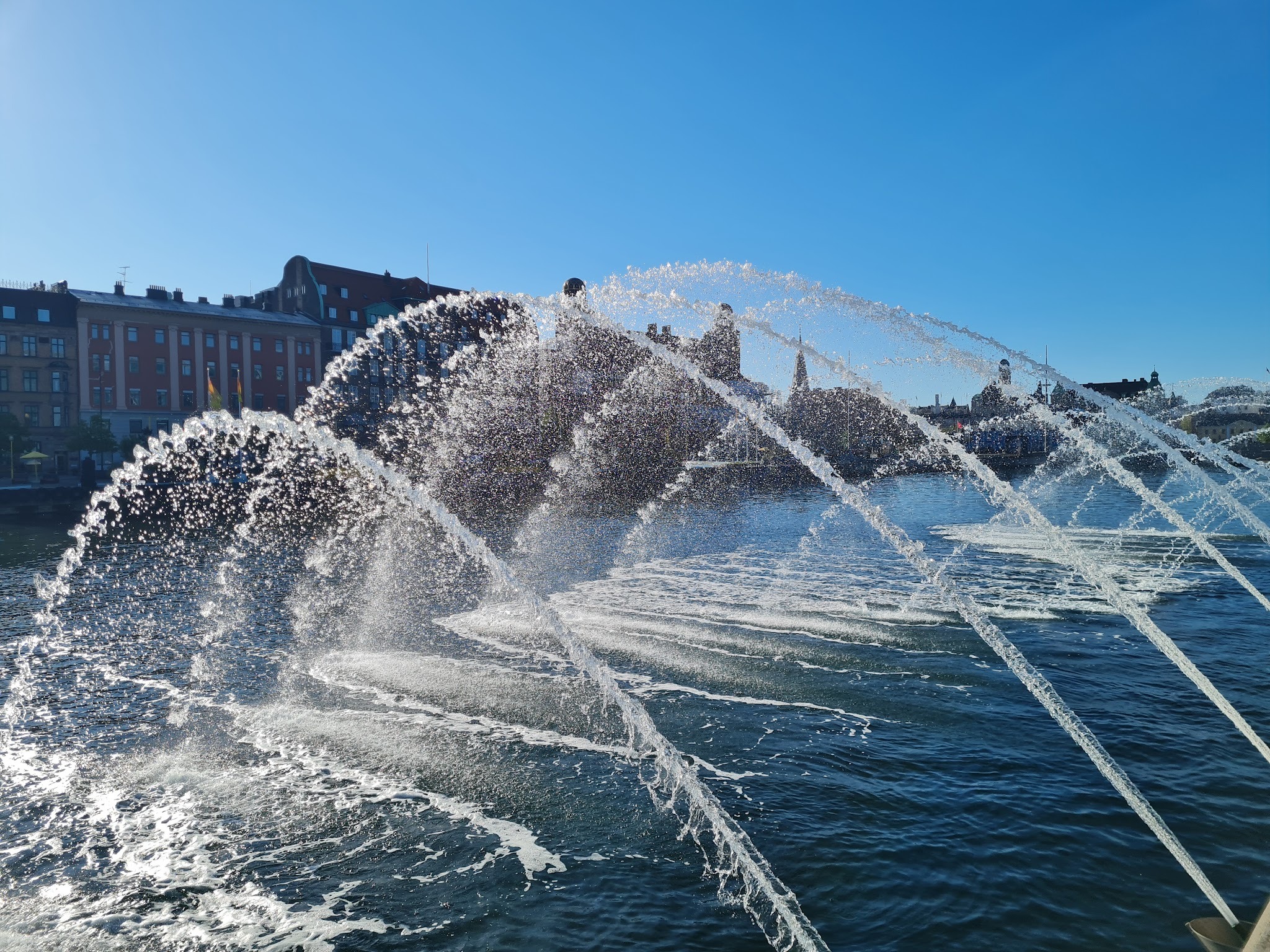 Sunlit water fountain spraying across Malmö harbor under a clear blue sky.