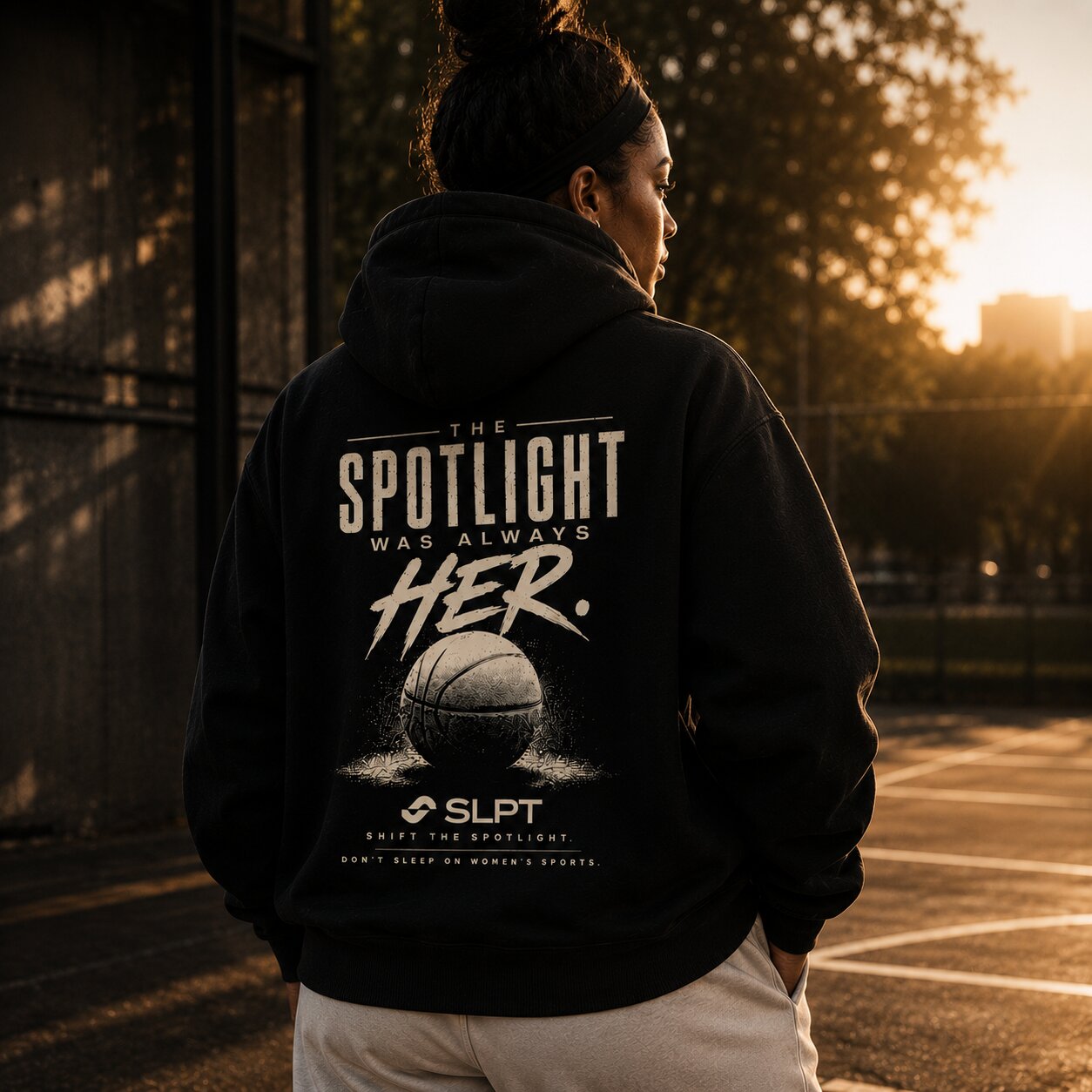 Person wearing SLPT hoodie on outdoor basketball court at sunset, featuring “The Spotlight Was Always Her” design.