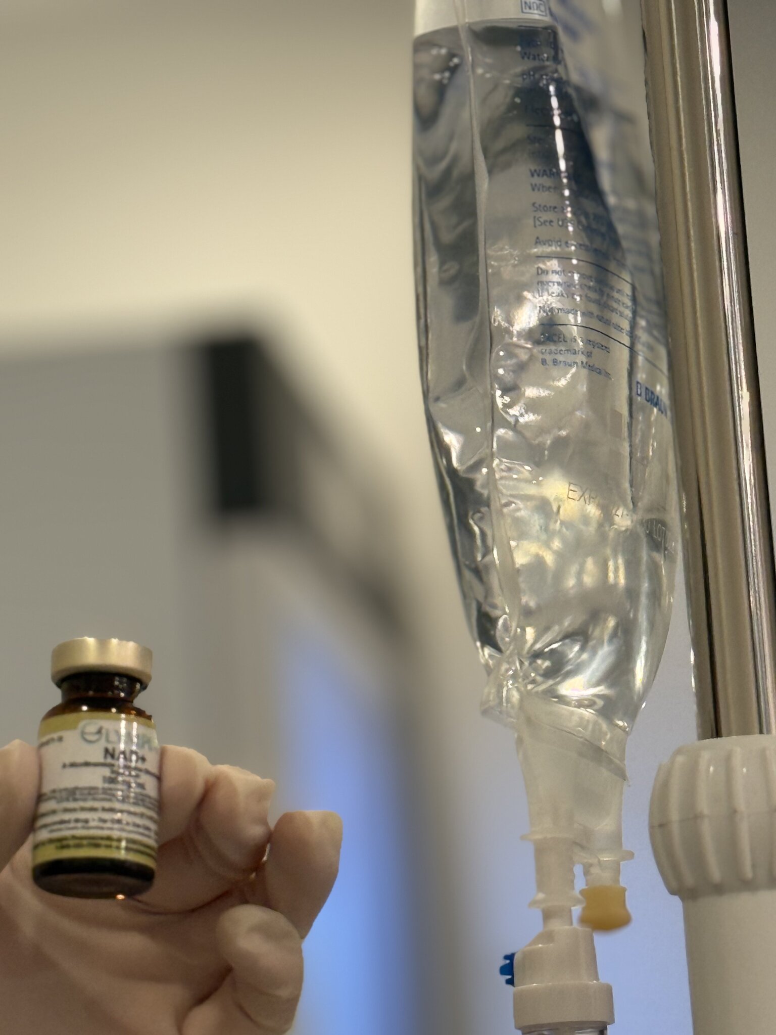 Gloved hand holding NAD+ vial next to IV infusion bag in treatment setting.