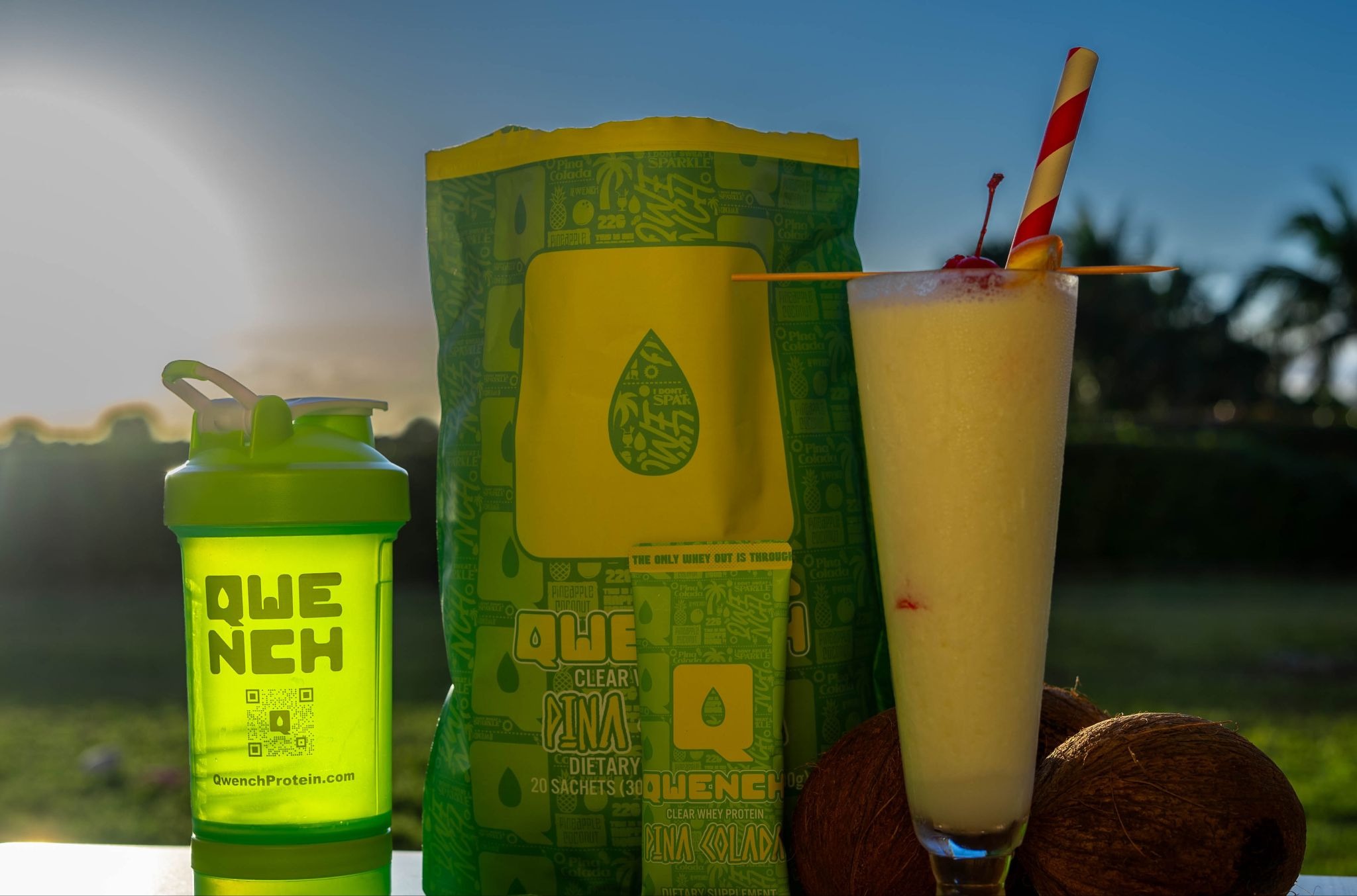 QWENCH Pina Colada protein pouch, shaker bottle, and tropical smoothie displayed outdoors at sunset.