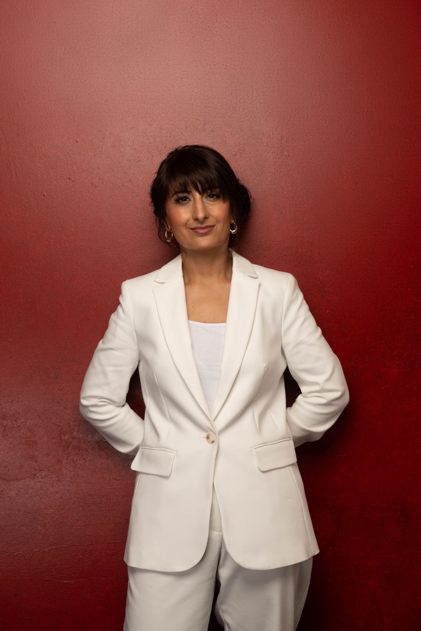 Kully Bath standing in a white suit against a textured red wall, smiling confidently.