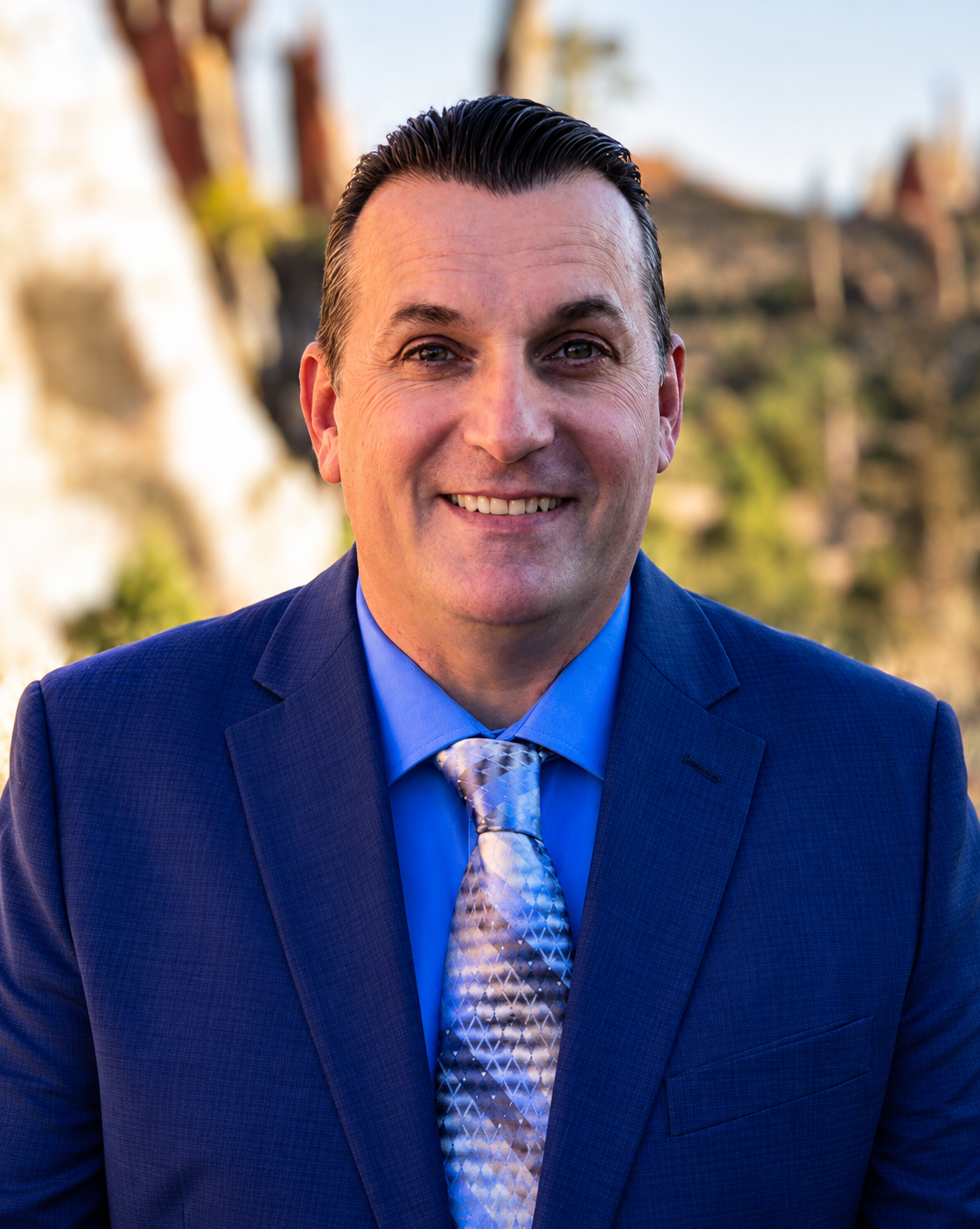 Professional headshot of a smiling man in a blue suit and patterned tie outdoors.