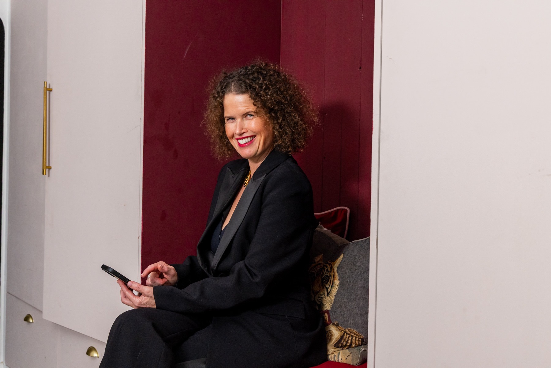 Dr Kate Armstrong smiling in a black suit, seated with a smartphone in a modern alcove.