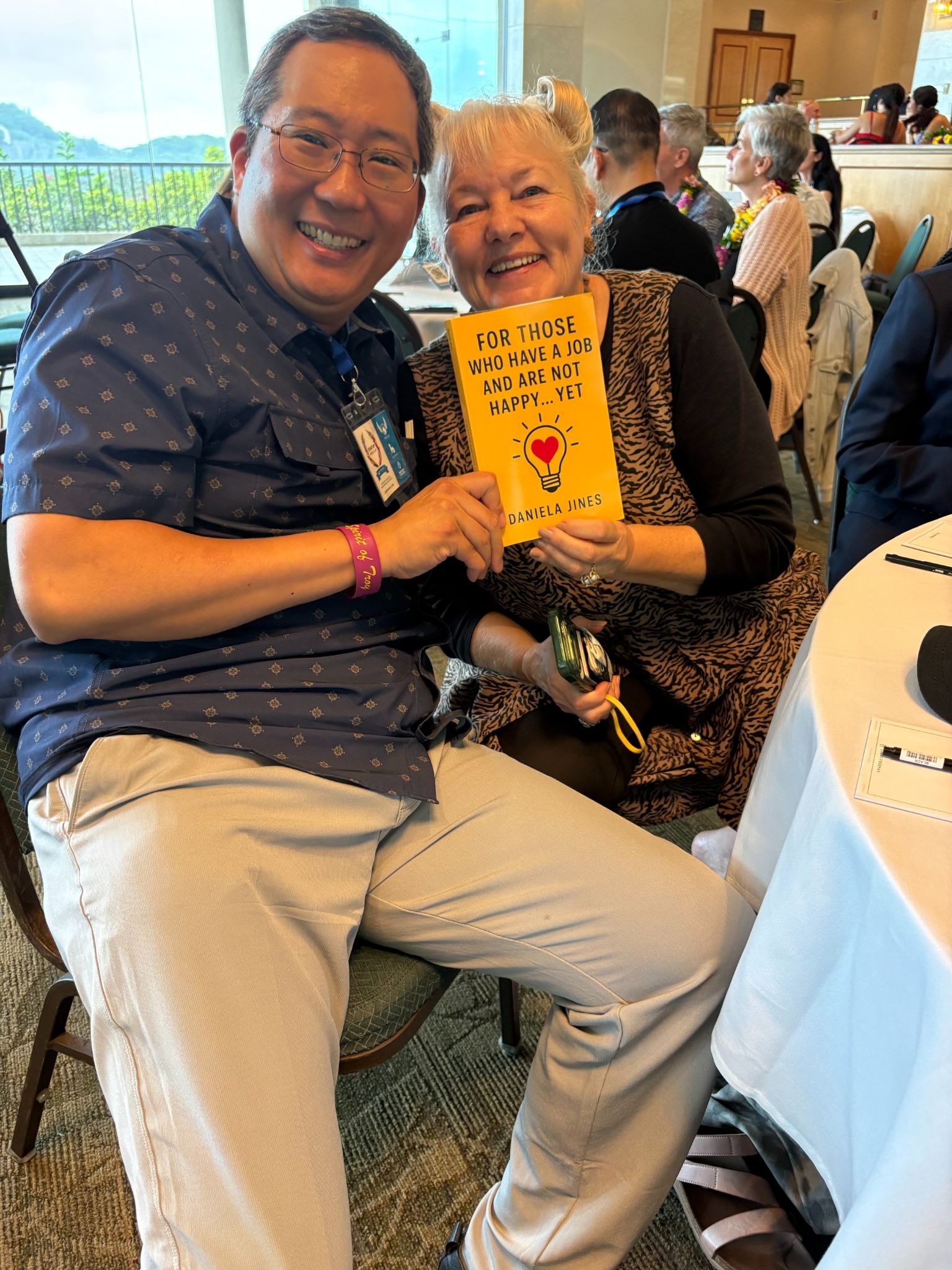 Two smiling attendees hold Daniela Jines' book at a networking event celebrating workplace happiness and leadership.