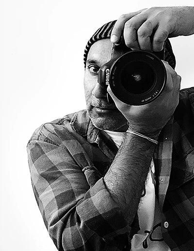 Black-and-white portrait of photographer Andrew Para holding a camera lens to his eye.