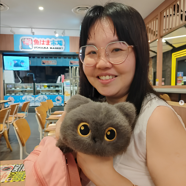 Woman smiling while holding a fluffy gray plush toy with large golden eyes in a café.