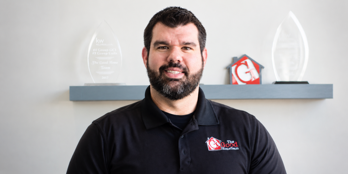 Nick Good wearing a black The Good Home Team polo with company awards displayed behind him.