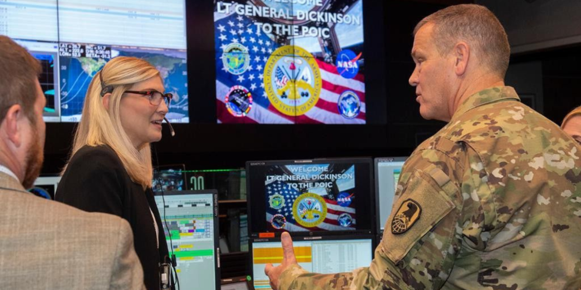 Military officer speaks with personnel inside a command center surrounded by computer monitors and U.S. military displays.
